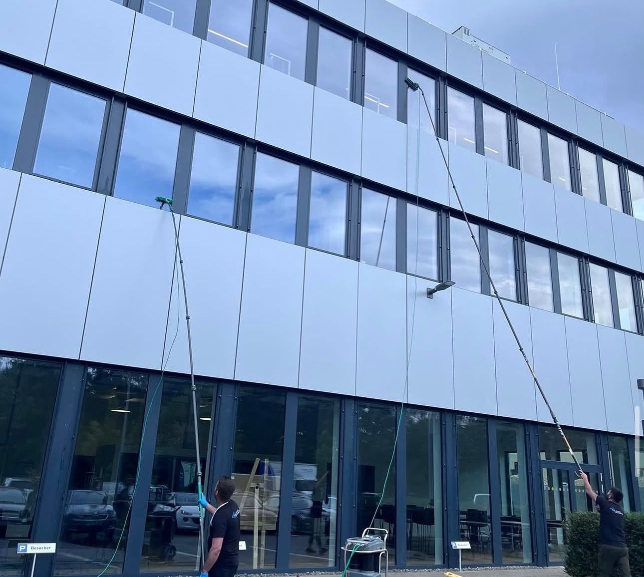 Zwei Mitarbeiter reinigen die Fenster eines Bürogebäudes mit Teleskopstangen vom Boden aus